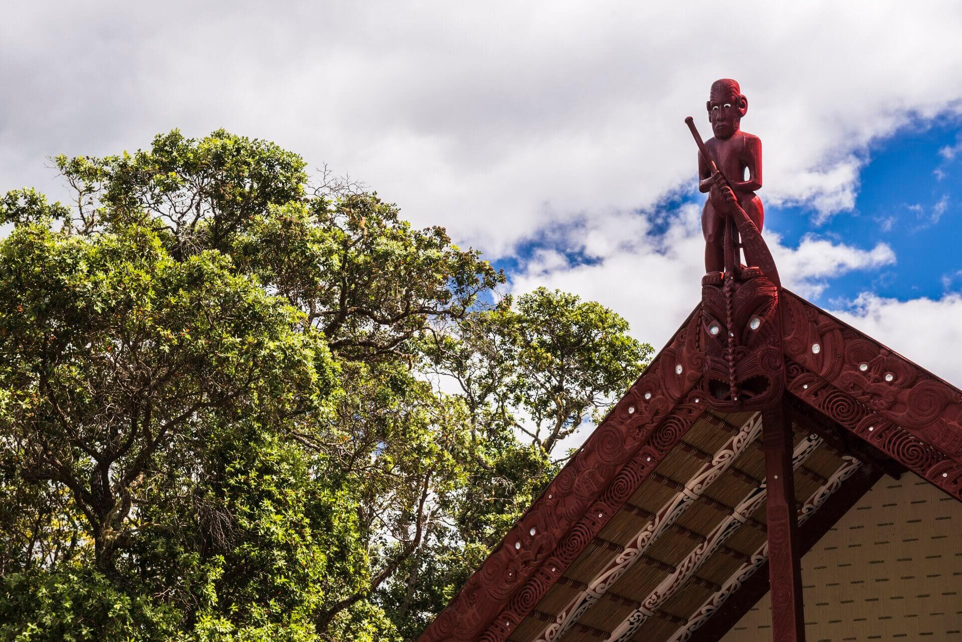 Traditional Māori meeting house with ornate red carved roof and ancestor figure, surrounded by native New Zealand forest beneath a partly cloudy sky