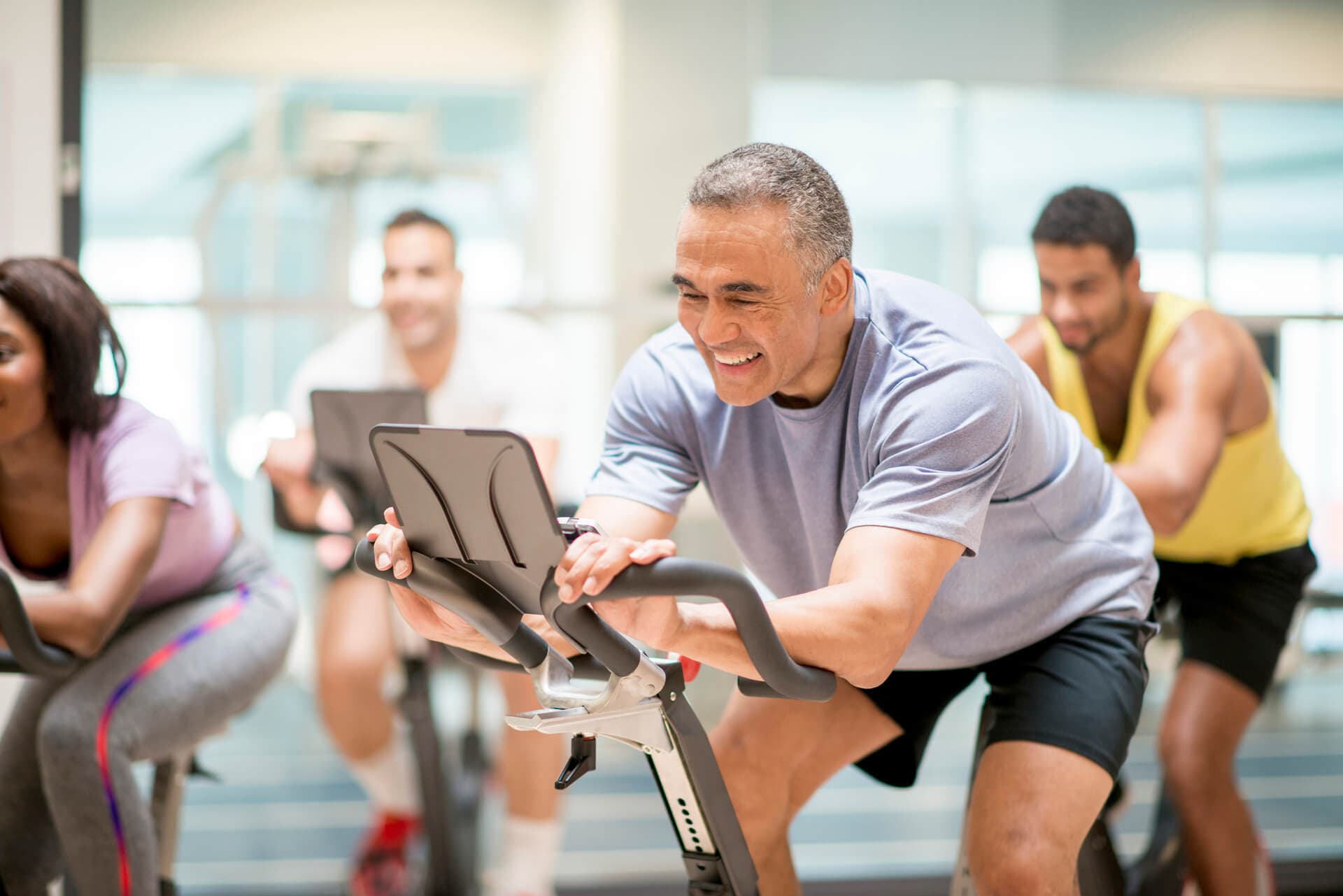 Older man smiles, riding an exercise bike in a class of other bikers